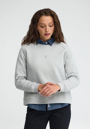 Mujer con cabello castaño ondulado que lleva una sudadera gris clara sobre una camisa vaquera azul, ajustándose las mangas sobre un fondo liso.