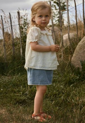 Fiatal lány fehér hímzett blúzban, farmer szoknyában és szandálban áll, kezében száraz fűvel egy füves kültéri területen, amelyet fa kerítés határol.