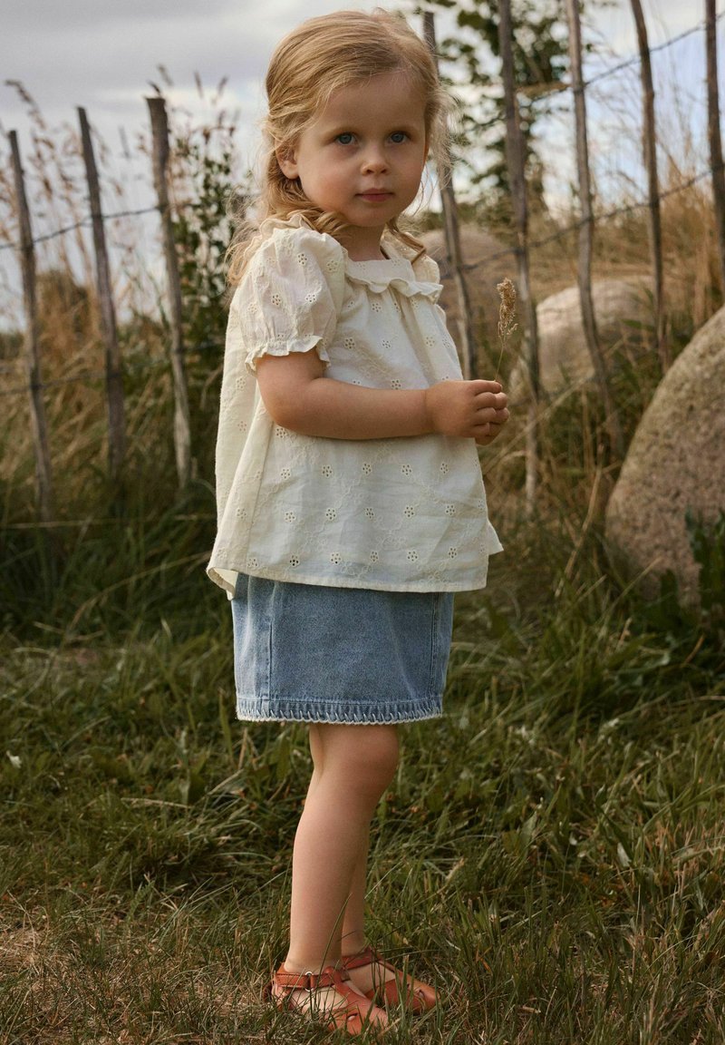 Jeune fille en blouse blanche à œillets, jupe en jean et sandales, debout tenant de l'herbe sèche dans un espace extérieur herbeux avec une clôture en bois.