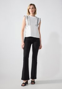White top with ruffled black accents at the shoulders, paired with flared black jeans and black sandals with knot detail.