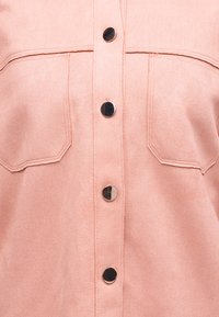 Minkštos rožinės spalvos marškiniai su dviem krūtinės kišenėmis ir keturiais blizgiais juodo metalo mygtukais priekyje centre.