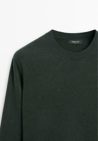 Donkergroen gebreide trui met een ronde hals, zachte textuur en ribbelrandjes. Etiket zichtbaar bij de hals geeft de merknaam aan.