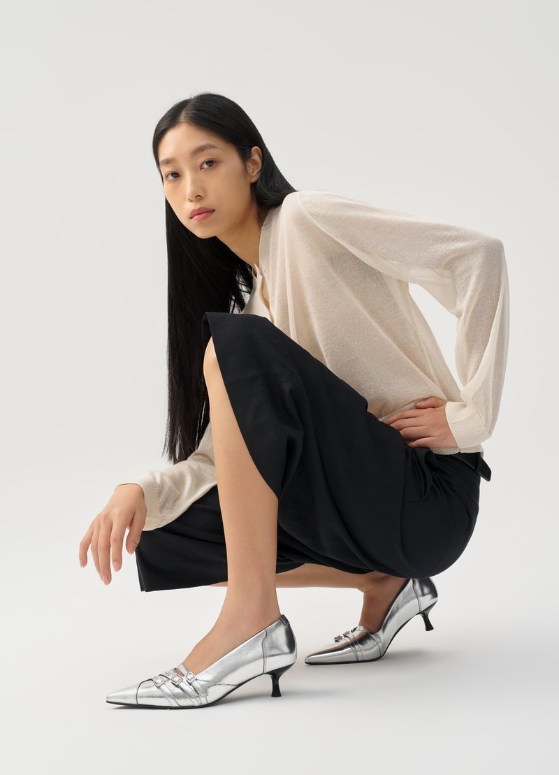 Donna con lunghi capelli neri che indossa un top beige trasparente, una gonna nera e scarpe basse argentate, accovacciata su uno sfondo bianco.