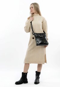 Abito maglione in cable knit beige, lunghezza al ginocchio con spacco laterale. Borsa a tracolla nera in pelle verniciata con finitura testurizzata e dettagli hardware. Stivaletti neri.