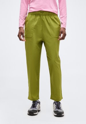 Persona que lleva pantalones de cintura elástica color verde oliva con bolsillos frontales, camisa de manga larga rosa y zapatos de senderismo gris y negro, de pie sobre un fondo blanco.