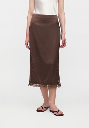 Femme portant une jupe midi en satin marron avec bordure en dentelle et des tongs noires, debout devant un fond clair uni.