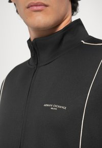 Chaqueta negra con cremallera fabricada en un tejido suave, con costuras de contraste claras y el logo bordado "Armani Exchange Milano" en el pecho.