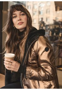 Chaqueta acolchada de bronce metálico con cuello y puños de piel sintética negra, combinada con un suéter de cuello alto negro. Sosteniendo un vaso de café de papel.
