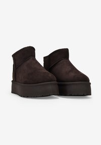 Botas de tobillo de ante marrón con puntera redondeada, suela plana y plataforma, y paneles laterales elásticos para facilitar su uso. El acabado texturizado añade detalle.
