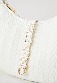 White handbag featuring a quilted texture and a gold hardware accent with the brand name "Valentino" embossed on it. Zippered closure.