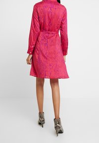 Abito fucsia con cintura, caratterizzato da un motivo a graniglia di legno, maniche lunghe e bottoni decorativi ai polsini. Indossato con tacchi stampa serpente.