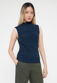 Top sin mangas de color marino con cuello alto, tejido texturizado y un ligero brillo, combinado con pantalones de pierna ancha en verde oliva, con las manos en los bolsillos.