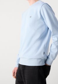 Sweatshirt bleu clair avec un col rond, des poignets et un ourlet côtelés. Présente un petit logo sur la poitrine et un détail sur la couture latérale.