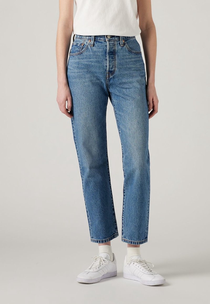 Personne portant un jean bleu taille haute, des baskets blanches, des chaussettes blanches et un top blanc sans manches rentré à l’intérieur, sur un fond uni clair.