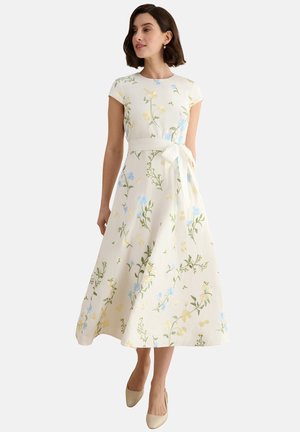 Frau trägt ein weißes Midikleid mit kurzen Ärmeln, Blumenmuster in Blau und Gelb und einem Taillengürtel, kombiniert mit beigen High Heels.
