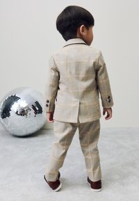 Enfant portant un costume léger à carreaux aux tons beige et jaune, composé d'une veste cintrée et d'un pantalon à plis plats. Chaussures marron à semelles blanches posées à côté.