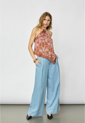 Kobieta w brązowym topie na szyję z wzorem, szerokich jasnoniebieskich dżinsach, czarnych szpiczastych butach i złotych bransoletkach, stojąca z rękami w kieszeniach.