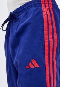 Niebieskie sportowe szorty z czerwonymi trzema paskami po bokach, elastycznym pasem z troczkiem i czerwonym trójkątnym logo przy kieszeni.