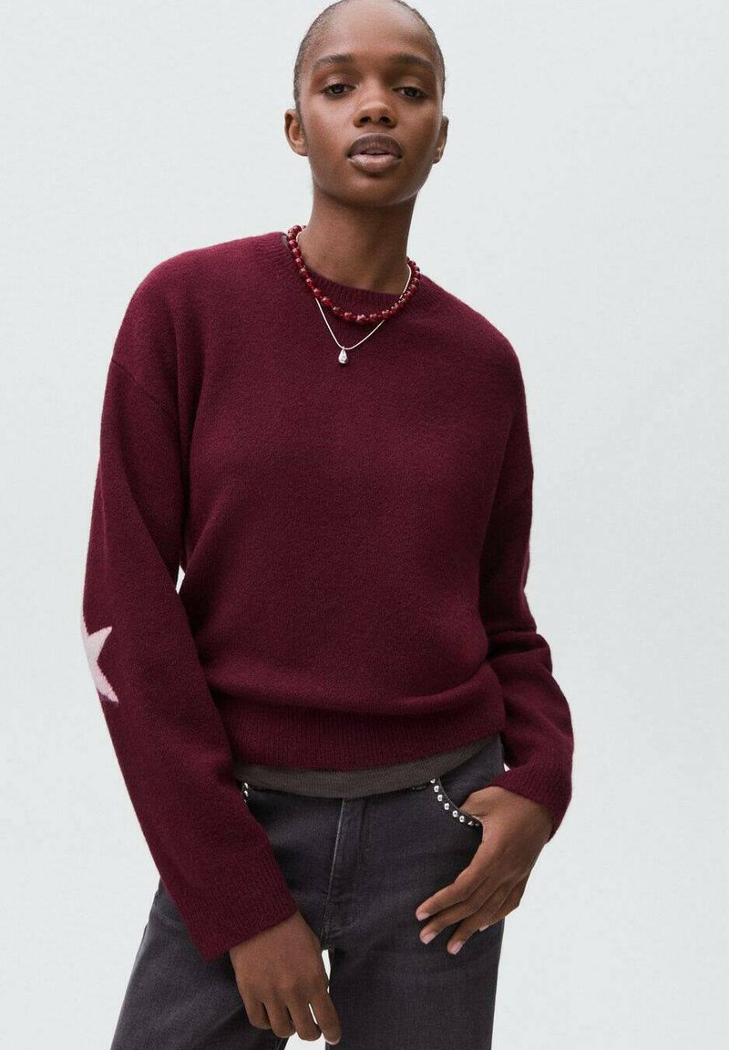 Eine Frau trägt einen bordeauxfarbenen Pullover mit einem hellen Stern auf dem Ärmel, über einem grauen Shirt, kombiniert mit schwarzen Hosen mit Nieten-Taschen.