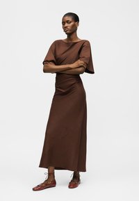 Γυναίκα που στέκεται με σταυρωμένα τα χέρια, φοράει καφέ τοπ με κοντά μανίκια και μακρύ φόρεμα σετ, με κόκκινα παπούτσια με κορδόνια.