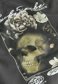 Tissu noir avec un graphique représentant un crâne, des fleurs et un papillon, détaillé avec des couleurs atténuées et un logo blanc en bas.