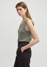 Soepele, olijfgroene mouwloze top met een diepe V-hals, gemaakt van gladde stof, gecombineerd met hoge, donkerbruine broeken.