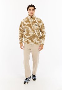 Fleece pullover i beige med hvide mønstre, kvart-zip lukning og frontlomme. Parret med lysebeige bukser og mønstrede sneakers.