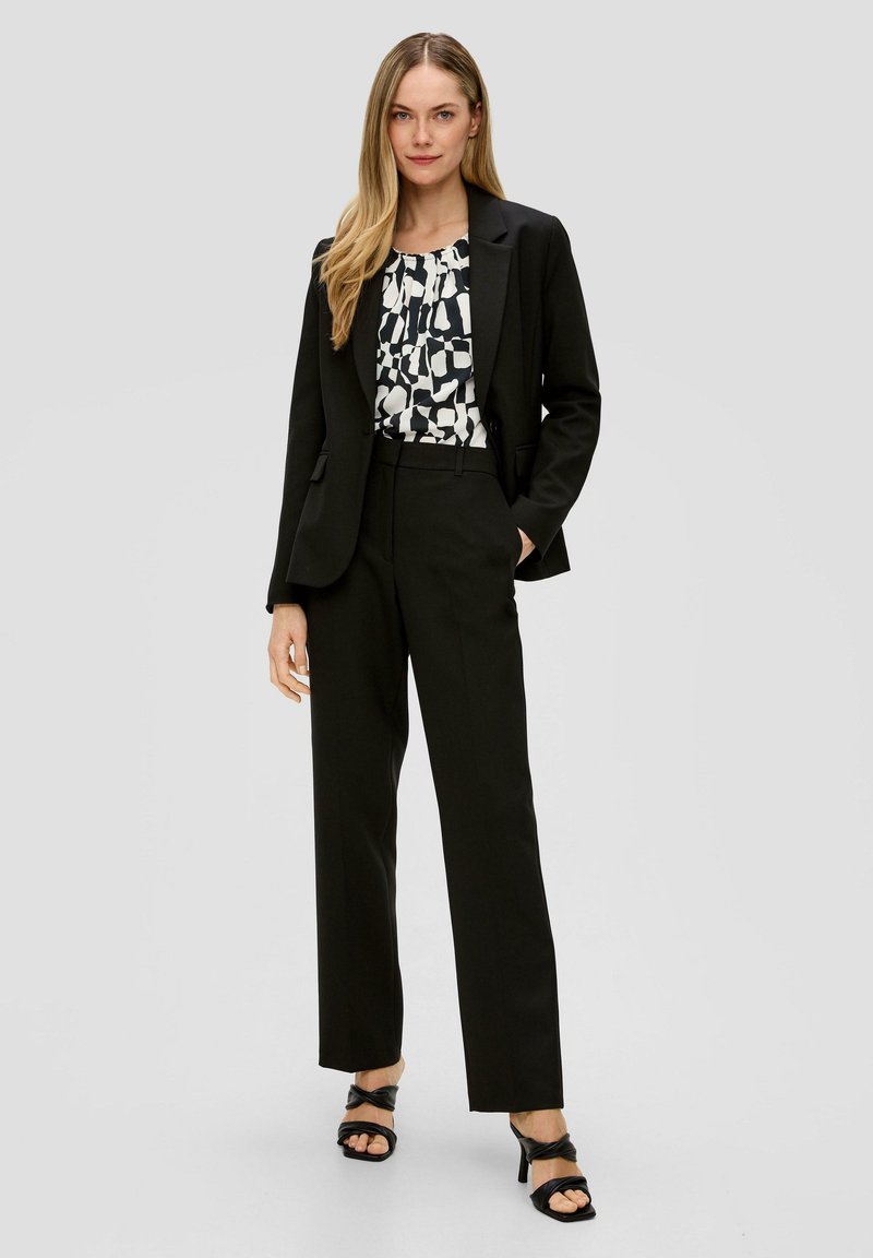 Zwart op maat gemaakt pak met een getailleerd blazer en wijde broekspijpen, gecombineerd met een gebloemde blouse in zwart en wit, en zwarte hakken.