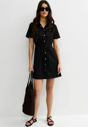 Vrouw draagt een zwart, met knopen voorzien, korte mouw jurk, zwarte zonnebril, zwarte sandalen, houdt een grote zwarte gevlochten shopper vast, staat tegen een witte muur.