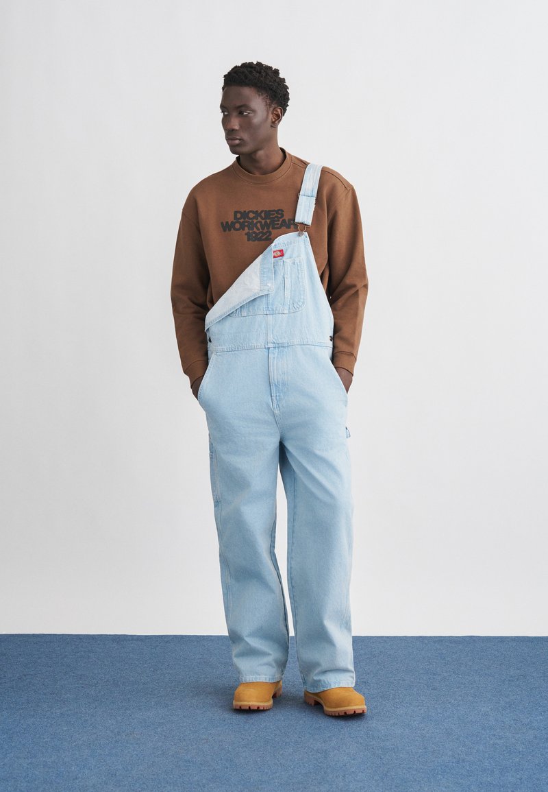 Mann trägt hellblaue Jeans-Latzhose mit einem losen Träger, braunen Pullover mit der Aufschrift "Dickies Workwear 1922" und beige Arbeitsstiefel.