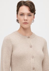 Gaišs adīts kardigans ar noapaļotu kakla izgriezumu, ar piecām pogām priekšpusē un mīkstu, teksturētu audumu.