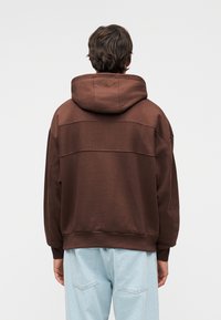 Καφέ φούτερ με φαρδιά κουκούλα, άνετη εφαρμογή και αντίθετα υφασμάτινα πάνελ· φορεμένο με ανοιχτόχρωμο μπλε τζιν παντελόνι.