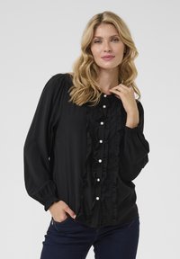 Blusa nera con rouches frontali e bottoni perlacei, maniche lunghe con polsini arricciati, tessuto testurizzato, design aderente, indossata con jeans scuri.