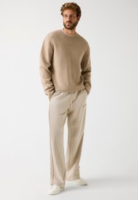 Felpa beige con logo ricamato, abbinata a pantaloni della tuta beige chiaro con strisce laterali. Entrambi realizzati in tessuto morbido. Vestibilità casual.