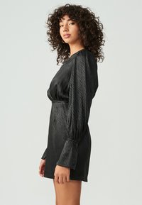 Zwarte jurk met gestructureerde stof, opgeblazen mouwen en een getailleerde silhouet. Bevat een geraffineerde taille en subtiele glans.