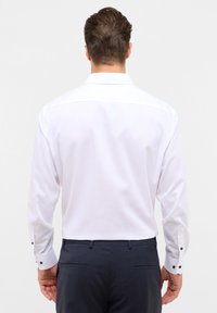 Camicia bianca a maniche lunghe con colletto formale, texture liscia e dettagli in bottone nero sui polsini. Indossata con pantaloni scuri, vista da dietro.