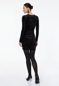 Frau in einem schwarzen samtigen Longsleeve-Minikleid, schwarzen Strumpfhosen, schwarzen High Heels und großen goldenen sternförmigen Ohrringen, die sich abgewandt zeigt.