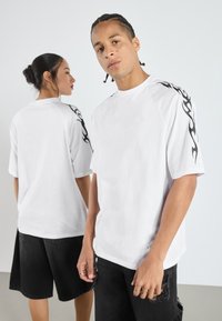 Camisetas blancas con diseños tribales negros en los hombros, combinadas con pantalones cortos negros, exhibidas por dos modelos que miran en direcciones opuestas.