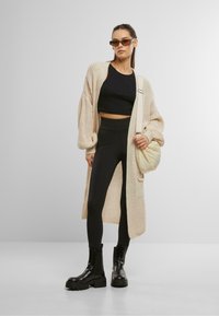 Chunky beiger Strickcardigan über einem schwarzen, verkürzten Oberteil und hochgeschnittenen schwarzen Leggings, kombiniert mit schwarzen Plateaustiefeln und einer gesteppten Tasche.