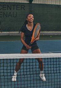 Tennis spiller i en svart t-skjorte med hvite detaljer, holder en oransje racket. Bane med blå overflate, nett i forgrunnen.