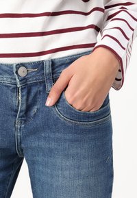 Jeans de mezclilla azul con un corte clásico, que cuentan con un bolsillo frontal y un botón plateado. Una parte de una camiseta a rayas blanca y burdeos es parcialmente visible.