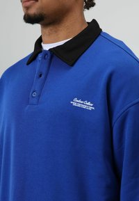 Blauwe pullover met een zwarte kraag, voorzien van een knoopsluiting met twee knopen en een wit geborduurd logo op de borst. Gladde stof, casual ontwerp.