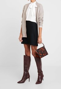 Cardigan beige a costine, blusa bianca con fiocco, gonna nera, stivali marroni sopra al ginocchio e una borsa a tracolla in tartaruga nera e marrone.