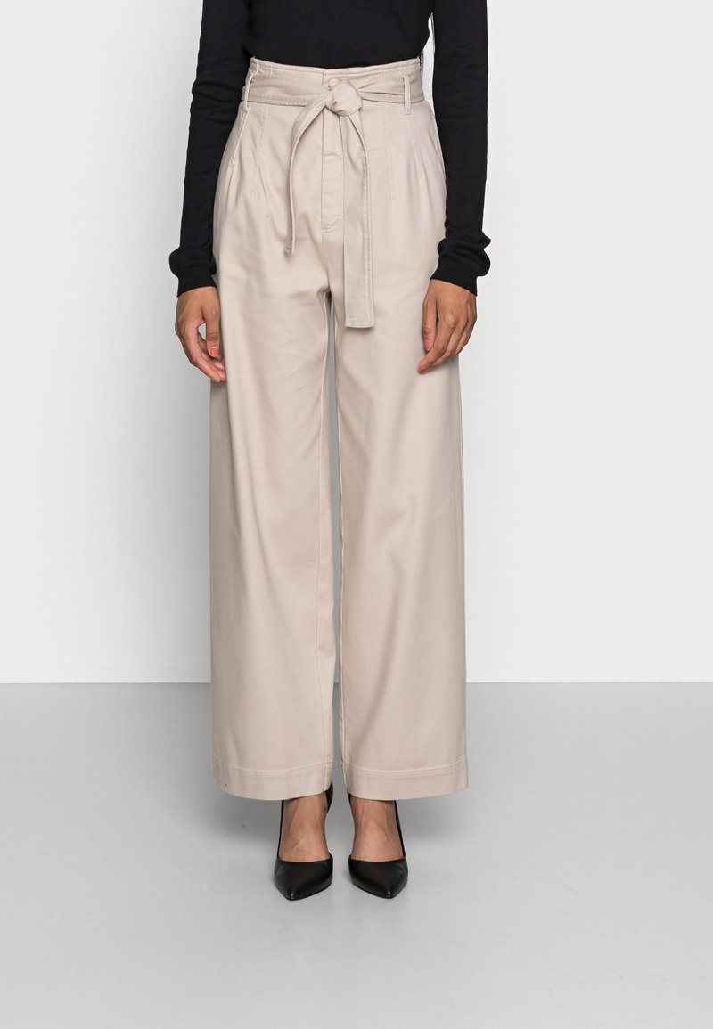Pantalons larges taille haute beiges avec une ceinture en tissu, présentant des plis et une texture lisse. Des escarpins noirs à bout pointu complètent la tenue.