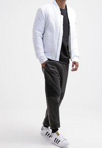 Uomo che indossa una giacca bomber bianca, una felpa nera, pantaloni jogger neri texturizzati e sneakers bianche Adidas, mentre cammina su uno sfondo bianco.