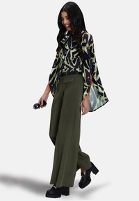 Blusa fluida multicolore con motivi astratti in verde e viola, abbinata a pantaloni larghi a vita alta verde oliva e scarpe nere massicce.
