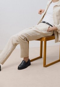 Personne portant un pantalon beige, une veste assortie, une chemise blanche et des chaussures noires assise avec les jambes étendues sur une chaise en bois simple.