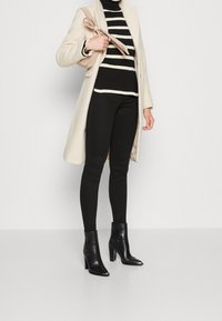 Beżowy wełniany płaszcz, czarno-biały sweter w paski, czarne skinny jeansy oraz czarne botki na obcasie z frędzlami.