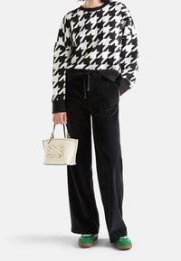 Person klädd i svart och vit houndstooth-tröja, svarta byxor, gröna skor, hållande en krämfärgad handväska med broderad design, stående mot vit bakgrund.