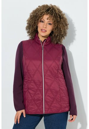 Donna con capelli ricci che indossa un gilet trapuntato bordeaux senza maniche sopra una maglia a maniche lunghe viola scuro e jeans blu, in piedi contro un muro bianco.